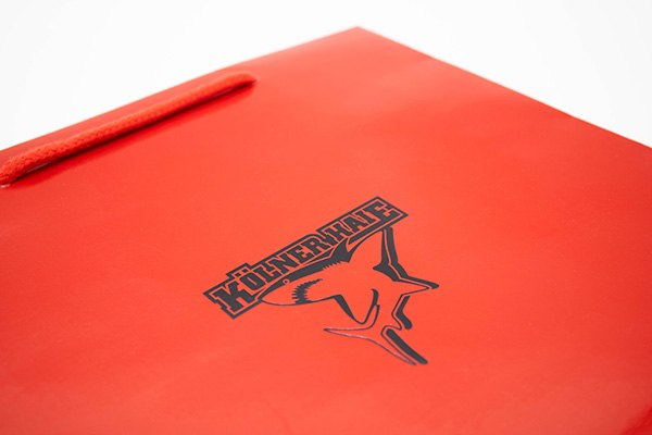 Rote Papiertragetasche mit schwarzem Kölner-Haie-Logo und Kordelgriff